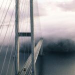 A dramatic view of a fog-covered suspension bridge with cables and the ocean below.